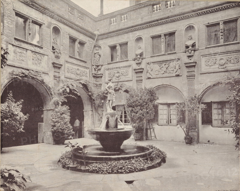Courtyard of the castle of Pierrefonds, album page, photomechanical print, This print is part of an album., maker: Angerer & Göschl, C., 1880 - 1900, paper, autotype, height 201 mm x width 248 mm, courtyard, castle, Pierrefonds, Castle of Pierrefonds, A stone courtyard with arched walkways and a two-story brick facade featuring ornamental relief panels and recessed niches holding sculpted busts and figures, a central multi-tiered fountain with a statue atop a pedestal and water spouts, surrounding low circular planting bed with foliage, potted shrubs and small trees placed near the walls, shuttered windows and doorways beneath the arches, climbing plants and vines on the masonry, and decorative balustrade and cornice details along the upper edge of the building.