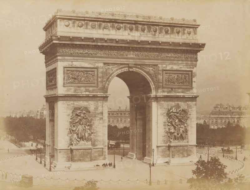 Arc de Triomphe in Paris, album page, photograph, This photo is part of an album., inscription: '50. Paris_ Arc de Triomphe', 1880 - 1910, photographer, paper, albumen print, height 214 mm x width 273 mm, Person, Napoleon I (Emperor of the French), triumphal arch, Paris, Arc de Triomphe, A large stone triumphal arch fills the frame, showing detailed sculptural reliefs on its pillars and above the central arched opening, with ornate decorative friezes along the top; the arch stands on a paved plaza bordered by rows of trees and flanked by multi-story buildings in the background, with horse-drawn carriages and small groups of people visible near the base of the monument; lamp posts and a low chain barrier line the plaza, and there are sculpted figures in high relief on the faces of the arch's piers.