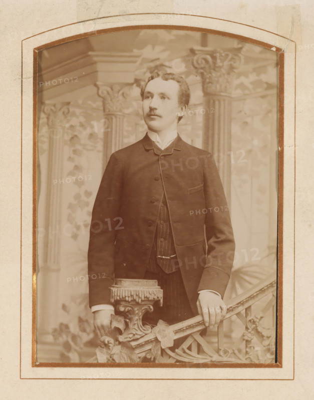 Portrait of a young man, standing by a stair railing, album page, photograph, cabinet photograph, This photo is part of an album., inscription: 'Frans E. Arvidsson, Helsingborg, Drottninggatan 18., Plåten förvaras, No... för efterbeställning.', photographer: Frans Edvard Arvidsson, 1872 - 1900, paper, pasteboard, albumen print, albumendruk, height 139 mm x width 98 mm, historical persons, standing figure, youth, adolescent, staircase, A sepia-toned portrait of a young man standing, shown from head to mid-thigh, wearing a dark suit jacket over a vest and a high-collared shirt; he has short wavy hair and a small mustache and looks slightly to his left, with one hand resting on an ornate carved railing that features decorative leaves and a fringed upholstered finial, and a painted backdrop behind him shows architectural columns and vine-like foliage; the photograph is mounted within a worn paper frame with rounded top corners and visible tape and discoloration along the edges.