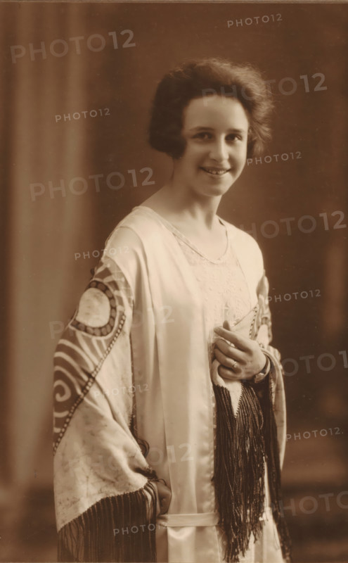 To Mr. E van Essen, Nora Janssens, Sept 1925 A'dam, Portrait of Nora Janssens, photograph, The photo is mounted on a passe-partout from photographer Geveke and placed in a folder., annotation: 'To Mr. E van Essen, Sept 1925 A'dam', signature: 'F. Geveke A'dam 10 Sarphatipark', photographer: Frits Geveke, Amsterdam, 1925, Frits Geveke, photographer, paper, gelatin silver print, developing gelatin silver print, height 173 mm x width 115 mm, height 332 mm x width 236 mm, Person, historical persons - BB - woman, Netherlands, A vintage sepia portrait of a smiling young woman with short wavy hair, wearing a light satin robe with patterned sleeves and long fringe, a lace undergarment visible at the neckline, and a ring on one finger as she lightly clasps the fabric at her chest, set against a plain softly blurred backdrop.