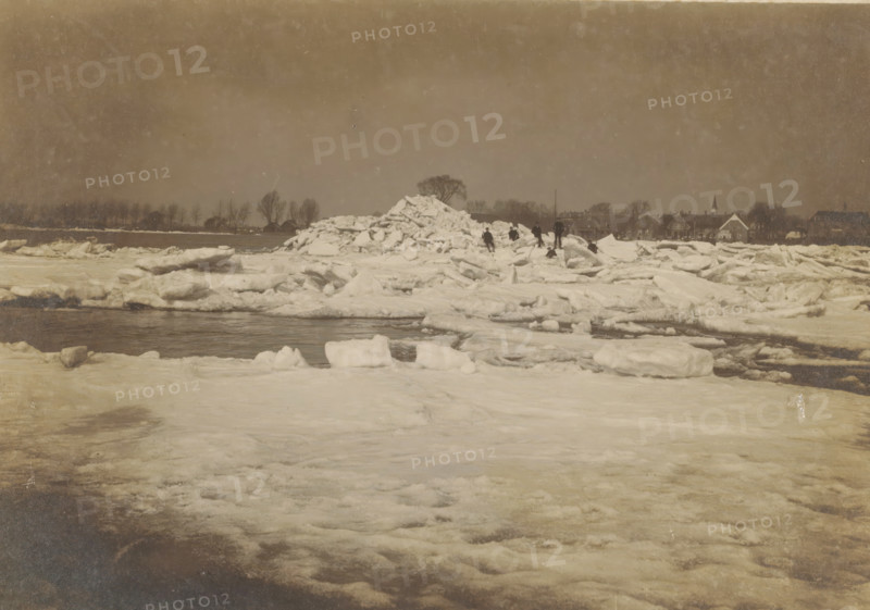 Ice floes in a river, Netherlands, photograph, check stamp: 'Atelier W. Visser Werkendam', photographer: W. Visser, Werkendam, 1910 - 1940, pasteboard, paper, gelatin silver print, developing gelatin silver print, height 120 mm x width 166 mm, height 239 mm x width 296 mm, river, drift ice, A wide frozen landscape with large sheets and chunks of ice piled and scattered across a snowy surface, a dark strip of open water cutting through the ice, several small figures standing and walking near a tall mound of broken ice, a distant shoreline lined with bare trees and a row of houses with pitched roofs and a steeple, a muted cloudy sky overhead.