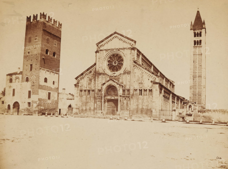 Verona. S. Zeno, View of the San Zeno in Verona, photograph, number: '644', inscription: 'n 8443, gel gr, part with pencil, indicated', Verona, 1851 - 1900, photographer, pasteboard, paper, albumen print, height 170 mm x width 229 mm, height 184 mm x width 234 mm, church (exterior), parts of church exterior and annexes: campanile, S. Zeno, A wide, sepia-toned photograph of a large stone church façade flanked by two tall towers, with the central façade featuring a large circular rose window above a carved arched doorway and vertical pilasters, the left tower constructed with rough brick and crenellations at the top adjacent to lower attached buildings with arched openings, and the right tower tall and slender with a pointed roof and patterned banding, all set against an open light ground in front and a pale sky above.