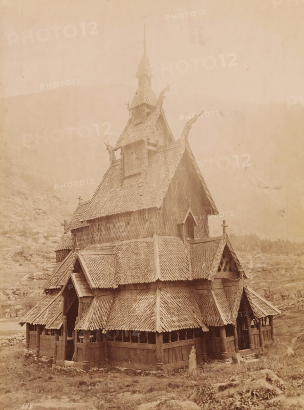 View of the Borgund Stave Church in Norway, photograph, inscription: 'Norge, Borgundskirke i Laerdal, Sogn.', Borgund, 1860 - 1900, photographer, Sogn og Fjordane, pasteboard, paper, albumen print, height 179 mm x width 134 mm, height 300 mm x width 245 mm, the Christian Churches, Borgund Stave Church, A multi-tiered wooden building with steep, shingled roofs and ornate carved roof ridges fills the frame, showing layered gables, small arched windows, and open arcaded walls supported by wooden posts; the upper tower rises to a pointed spire with additional small roofed openings and carved finials, while the lower sections display overlapping shingle patterns, entrance porches beneath pitched roofs, and weathered wooden surfaces; the structure sits on a grassy, rocky landscape with faint hills visible in the background under a pale sky.