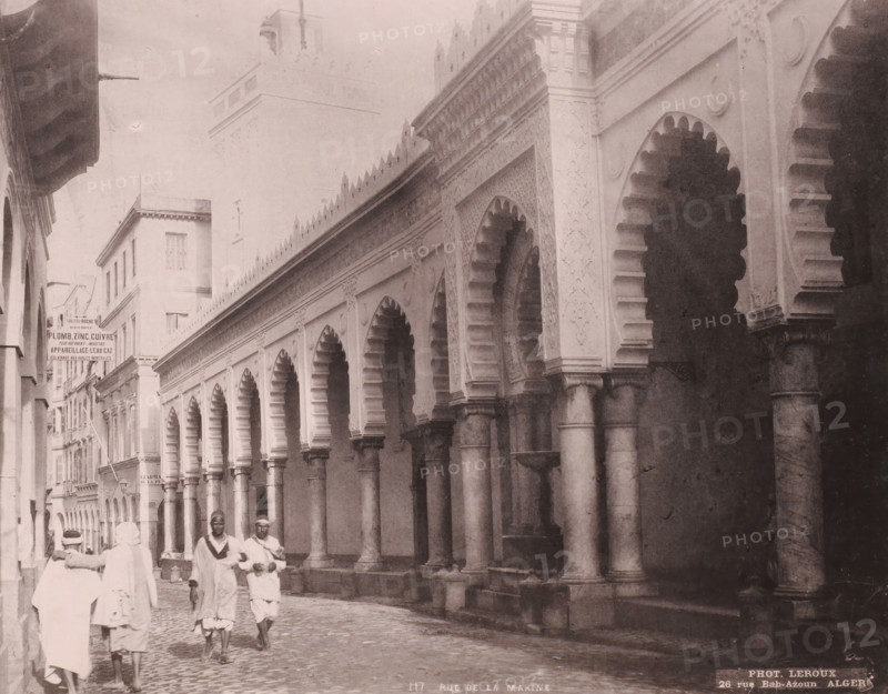 Rue de la Marine, View of the Djama'a al-Kebir in Algiers, photomechanical print, number: '117', address: '26 rue Bab-Azoun ALGER', photographer: Alexandre Leroux, Algiers, cliché maker: anonymous, c. 1875 - c. 1900, Alexandre Leroux, photographer, cliché maker, North Africa, pasteboard, paper, collotype, photoprint, height 247 mm x width 316 mm, temple, shrine, Islam, Mohammedanism, Djama'a al-Kebir, A narrow street scene with a row of tall, ornate arches supported by columns along one side of the street, each arch featuring scalloped inner edges and decorative panels above, and a series of buildings with multiple windows lining the opposite side; the street surface is cobbled and a small group of people wearing light-colored, loose garments and head coverings are walking and standing near the foreground, while the architectural details include carved motifs along the architrave, rounded column capitals, and a taller building or tower rising in the background.