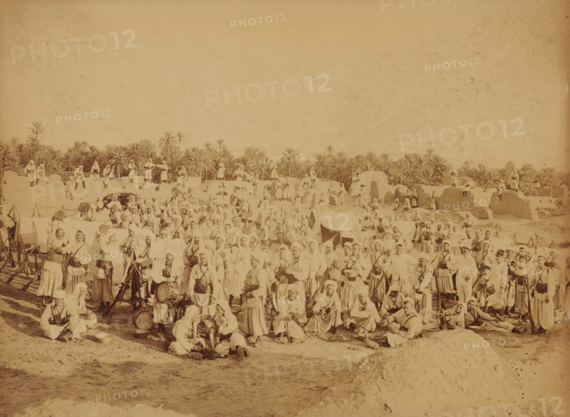 Fort Turc au Vieux Biskra, French garrison at Biskra, Algeria, Souvenir ... Biskra, photograph, inscription: 'Jul D...', Biskra, 1860 - 1900, photographer, North Africa, pasteboard, paper, albumen print, albumendruk, height 182 mm x width 241 mm, garrison, A wide sepia photograph showing a large group of people gathered outdoors with palm trees and low mud structures in the background; many individuals wear light-colored robes and head coverings, some hold musical instruments or drums, and others carry rifles or stand beside tripods and equipment; tents and makeshift shelters are scattered among the crowd, with people seated, standing, and leaning on the ground across the foreground and middle distance; the scene extends horizontally with figures positioned on earthen mounds and walls, and taller palm trees rising behind the settlement under an empty sky.