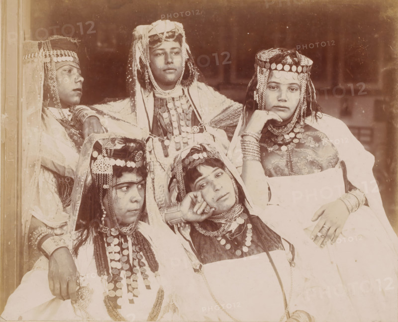 Group portrait of five unknown Ouled Naïl women in Algeria, Alger, groupe de filles d'Ouled Nail à Biskra, photograph, Algeria, 1890 - 1930, photographer, North Africa, paper, pasteboard, baryta paper, daylight gelatin silver print, height 247 mm x width 317 mm, anonymous historical persons depicted in a group, in a group-portrait - BB - woman, Five seated individuals wearing layered garments and headpieces decorated with round metal discs and dangling chains are shown in a close group portrait; some head coverings include veils and braided or beaded adornments, and several necklaces and bracelets with similar round metal decorations are visible on their chests and wrists; one person rests a hand under their chin while another leans their head on a hand, and their faces display neutral expressions against a plain, darker background.