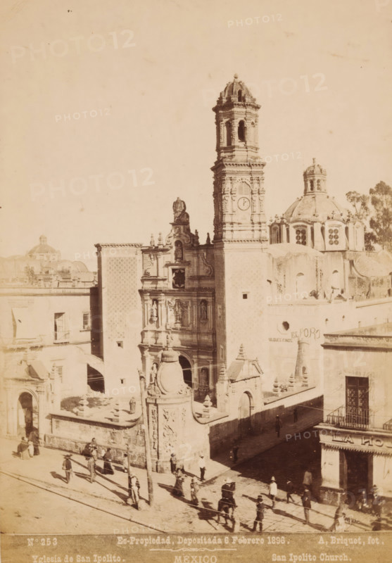 View of the San Hipólito y Casiano in Mexico City, Yglesia de San Ipolito, San Ipolito Church, Mexico, photograph, number: 'N. 253', copyright notice: 'Es-Propriedad, Depositada 6 Febrero 1896.', photographer: Abel Briquet, Mexico City, 1896, North America, pasteboard, paper, albumen print, height 193 mm x width 133 mm, height 210 mm x width 160 mm, Roman Catholic Church, San Hipólito y Casiano, A sepia-toned photograph shows an ornate stone church facade with a tall bell tower and a domed roof section; the facade features sculpted figures, decorative reliefs, and architectural details around a central entrance, with a carved columned gate and balustrade in front. People in period clothing are walking and standing along a sunlit street and near the church entrance, casting long shadows. Adjacent buildings with windows and balconies flank the church, and a narrow shaded alley runs beside it. Trees appear behind the domed roof, and the sky above is empty and pale.