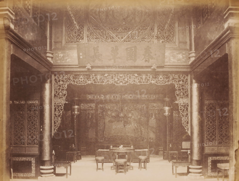 Interior of a Chinese house, photograph, annotation: 'Canton-Entrance to a Chinese House', 1850 - 1900, photographer, paper, pasteboard, albumen print, height 241 mm x width 310 mm, interior of the house, A sepia-toned interior scene showing an ornate room with carved wooden panels and decorative latticework framing a central area. Two large columns flank the foreground, each with visible bases and decorative bands. A horizontal plaque with large characters is mounted above an intricately carved frieze. Hanging lanterns descend on either side of the central space. In the middle of the room a round table sits on a low platform, surrounded by small carved stools and low chairs. Behind the table a large carved screen or panel features a central rectangular design with surrounding decorative borders. Additional small tables and stools are arranged along the sides of the room. The floor appears smooth and light in tone, and the overall scene contains repeating geometric and floral carved mo...