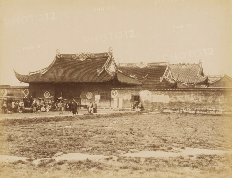 View of a city wall and residences in Shanghai, photograph, annotation: 'Shanghai-Chinese residences', Shanghai, 1850 - 1900, photographer, paper, pasteboard, albumen print, height 235 mm x width 310 mm, city-walls (city(-scape) with figures, staffage), A wide sepia photograph shows a long traditional building with sweeping tiled roofs and ornate upturned eaves, decorative ridge ornaments, and circular reliefs on the upper wall; a low wall runs along the front with a small doorway where a person stands partially sheltered by an umbrella, and a group of people gathered near the left side include figures standing, sitting, and carrying baskets and umbrellas; in front of the building a grassy open area stretches across the foreground.