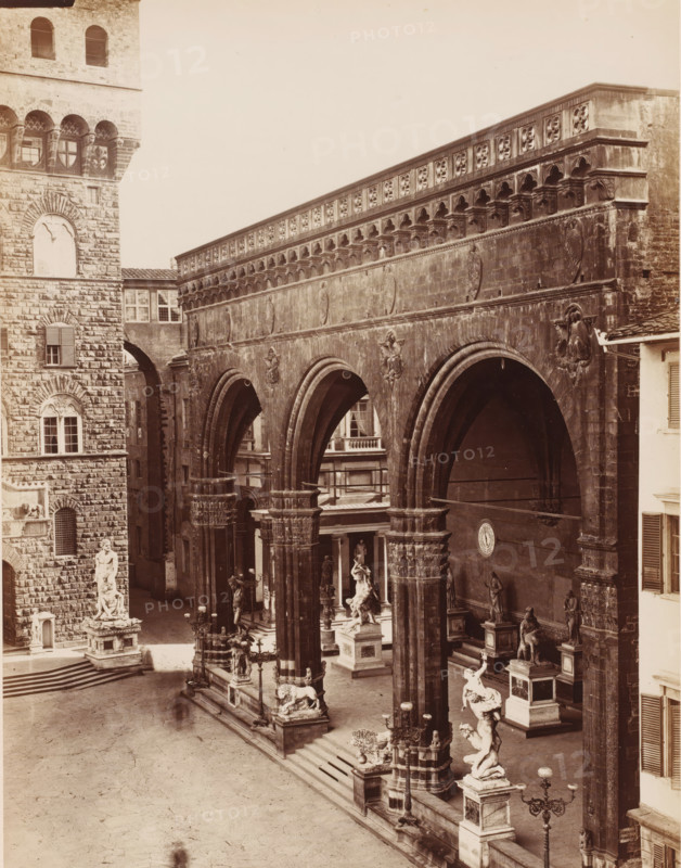 Loggia dell'Orgagna, View of the Loggia dei Lanzi in Florence, Italy, Firenze, photograph, number: '3057.', address: 'GIACOMO BROGI, PHOTOGRAPHEDITORE, FIRENZE, LUNG ARNO DELLE GRAZIE 15', photographer: Giacomo Brogi, Florence, publisher: Edizioni Brogi, 1864 - 1881, Giacomo Brogi, Edizioni Brogi, photographer, publisher, Tuscany, pasteboard, paper, albumen print, height 470 mm x width 344 mm, gallery of sculptures, Loggia dei Lanzi, A sepia-toned photograph shows a large stone arcade with tall rounded arches supported by ornate columns, decorative friezes along the upper wall, and sculpted reliefs on the façade; beneath the arches and along the elevated platform are multiple statues on plinths, including dynamic figures posed with limbs raised and seated figures, ornamental lamp posts, and animal sculptures; to the left is a textured stone building with arched windows and a tall sculpted statue on a pedestal at the base of steps leading into the open plaza; stairways and low balustrad...