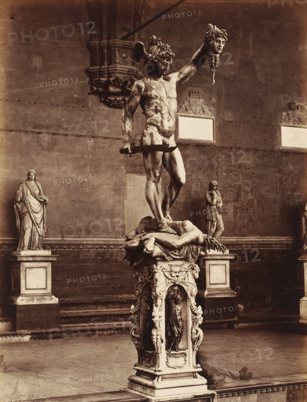 Sculpture Perseus by Benvenuto Cellini in the Loggia de Lanzi in Florence, Italy, Il Perseo del Cellini, Firenze, photograph, number: '3087.', address: 'GIACOMO BROGI, FOTOGRAFO-EDITORE, FIRENZE, ', photographer: Giacomo Brogi, Florence, after sculpture by Benvenuto Cellini, 1856 - 1881, Benvenuto Cellini, after sculpture by, Tuscany, pasteboard, paper, albumen print, height 469 mm x width 351 mm, piece of sculpture, reproduction of a piece of sculpture, Toscane, Loggia dei Lanzi, A large bronze statue of a muscular male figure stands on an ornate pedestal, the figure holding a severed head aloft in one hand and a curved sword in the other, a draped, reclining body lies beneath his feet on the pedestal, the pedestal is highly decorated with sculpted figures and ornamental motifs, a tall stone wall with plaques and architectural reliefs forms the background, two additional standing statues on plinths flank the central sculpture at either side, the scene is rendered in a sepia photograph...
