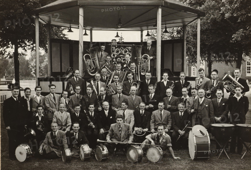 Group portrait of the 'Crescendo' harmony orchestra, photograph, photographer: NV Vereenigde Fotobureaux, Amsterdam, 1949, paper, gelatin silver print, gelatin silver print on photographic paper, height 120 mm x width 174 mm, anonymous historical persons portrayed in a group, in a group-portrait, banner, standard (as symbol of the state, etc.), musical instrument, group portrait, A large group of men posed together in front of a wooden bandstand, many wearing suits and ties and some in jackets, arranged in rows with brass and woodwind instruments visible throughout; several tubas and horns are positioned behind and beside the group, while trumpets, trombones, clarinets, and saxophones are held by seated and standing members; a row of drummers with snare drums and a bass drum sits at the front, with drumsticks and a drum major’s mace visible; the bandstand has a pitched roof supported by posts and hanging lamps, and decorative panels at the rear of the platform; trees and parkland are v...
