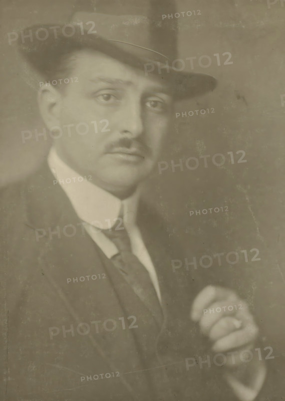 Portrait of the photographer Jacob Merkelbach, photograph, name: 'Bern. F.Eilers Amsterdam', photographer: Bernard Eilers, Amsterdam, 1910 - 1930, paper, daylight collodion silver print, height 230 mm x width 171 mm, height 407 mm x width 306 mm, Person, Jacob Merkelbach, portrait, self-portrait of photographer, A sepia-toned portrait of a man wearing a dark fedora and a suit jacket with a light-colored shirt and a tie, his face turned slightly toward the camera showing a thin mustache and a contemplative expression, one hand visible in the lower right holding an object, with a softly textured background and visible surface wear on the photograph.
