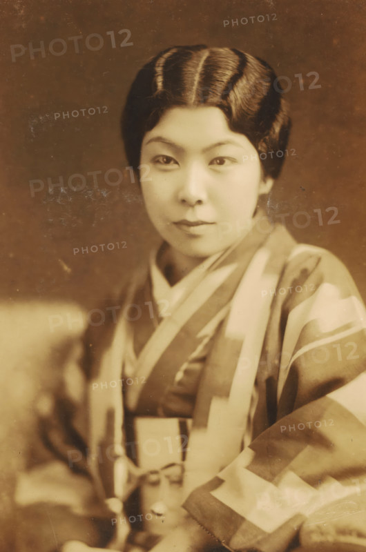 Portrait of an unknown Japanese woman, photograph, address: 'YOKOYAMACHO.HACHOJI., TEL. 320.', photographer: T Jingu, Tokyo, c. 1900 - c. 1920, Kanto, paper, pasteboard, baryta paper, daylight gelatin silver print, height 133 mm x width 89 mm, height 242 mm x width 156 mm, adult woman, anonymous historical person depicted, Japan, A sepia-toned portrait of a person wearing a patterned garment with broad stripes and geometric shapes, showing a close-up of the upper body and face. The person has neatly styled, dark hair with visible waves and a center part, and a smooth, even complexion. The eyes are looking forward, the eyebrows are softly curved, and the lips are closed. The garment has layered collars and draped fabric across the shoulders and chest, with a tied sash visible at the waist. The background is plain and softly textured, and the image has small surface marks and scratches.