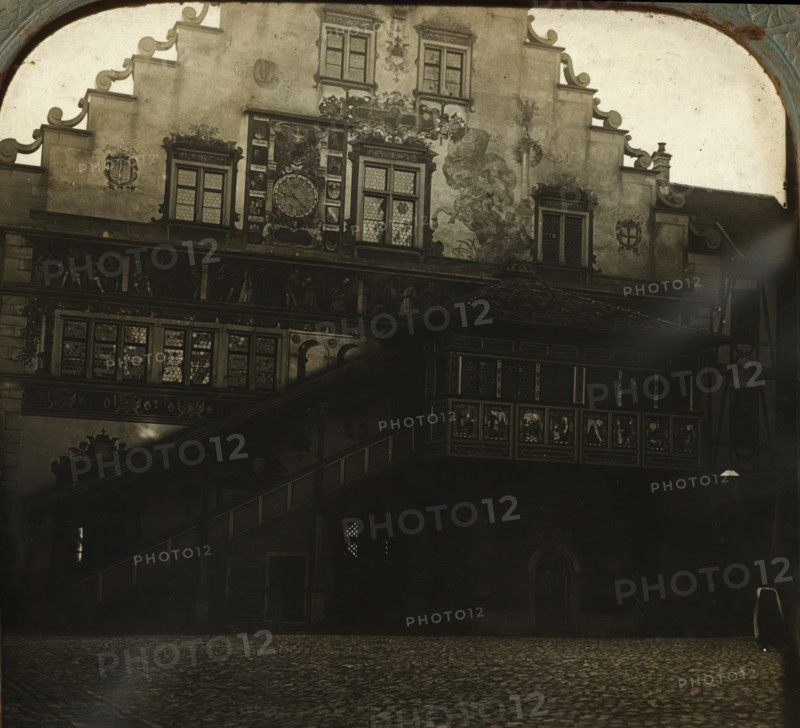 Rear facade of the Lindau town hall, Rathhaus in Lindau, Hinterseite, stereograph, number: '759.', Lindau im Bodensee, 1900 - 1940, photographer, Bavaria, paper, plastic, pasteboard, organic material, cellulose nitrate film, gelatin silver print, developing gelatin silver print, height 75 mm x width 150 mm, town hall, rear facade, A monochrome view of an ornate building façade with decorative gables and patterned windows, featuring a large circular clock framed by sculpted reliefs and coats of arms, painted murals or frescoes on the plastered wall, a projecting wooden gallery with carved panels and small leaded glass panes, an external staircase with a balustrade leading to the upper level, a cobbled foreground, and an arched decorative border at the top and sides of the image.