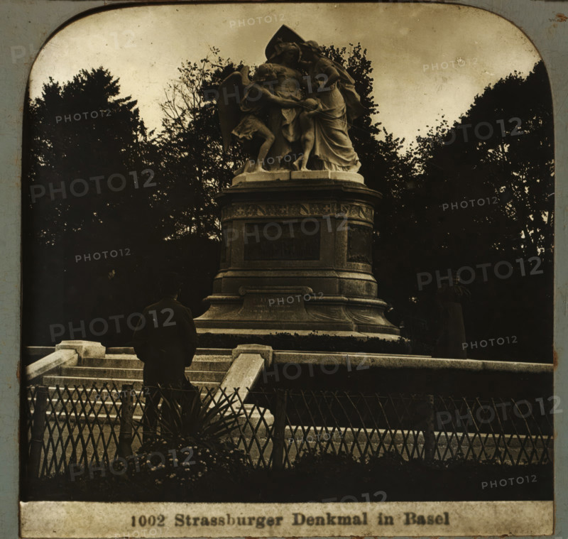 Strassburger monument in Basel, Strassburger Denkmal in Basel, stereograph, number: '1002', Basel, after sculpture by Frédéric Auguste Bartholdi, 1900 - 1940, Frédéric Auguste Bartholdi, photographer, after sculpture by, paper, plastic, pasteboard, organic material, cellulose nitrate film, gelatin silver print, developing gelatin silver print, height 75 mm x width 150 mm, monument, statue, A stone monument with a sculpted group on top stands amid trees and dark foliage, the sculpture depicting draped figures in flowing garments and wings on a raised pedestal with carved panels and decorative molding; stone steps and a low railing lead up to the base where a person in dark clothing faces the monument and another person stands to the side; a decorative fence and plants occupy the foreground and the sky above is pale.