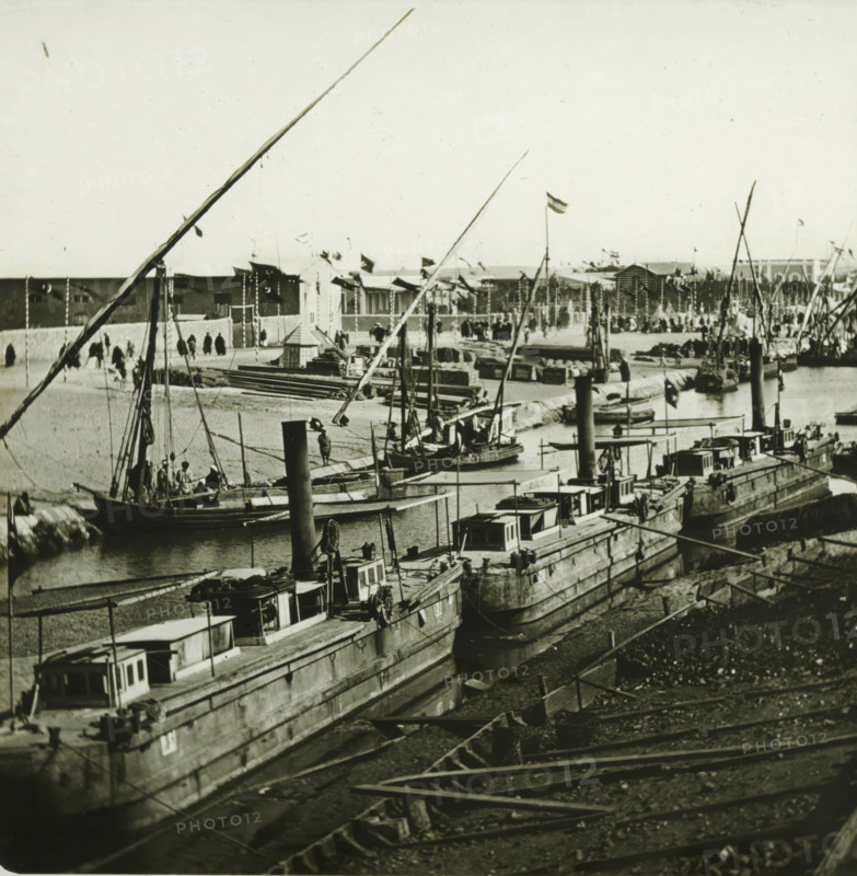 Inauguration of the Suez Canal. Ismaïlia, canal d'eau douce, arrival of the scheiks arabes (instantanee), Arrival of the Arab sheikhs for the opening of the Suez Canal, Egypt, stereoscopic photograph, number: '10011', photographer: Ferrier Père-Fils et Soulier & Léon et Lévy, Suez Canal, 1869, Ferrier Père-Fils et Soulier & Léon et Lévy, photographer, North Africa, inorganic material, glass, slide, height 84 mm x width 171 mm, canal, A waterfront scene showing multiple moored boats with tall masts and rigging alongside a quay, wooden decks and small cabin structures on the boats, and piles of cargo and crates on the shore; a row of larger vessels extends into the distance with visible chimneys and winches, and smaller rowing boats are tied near the waterline; long spars and ropes cross the foreground and middle ground, and people are visible on some boats and walking along the quay; beyond the quay a low wall separates the water from an open area with stacked materials, tents or tempor...