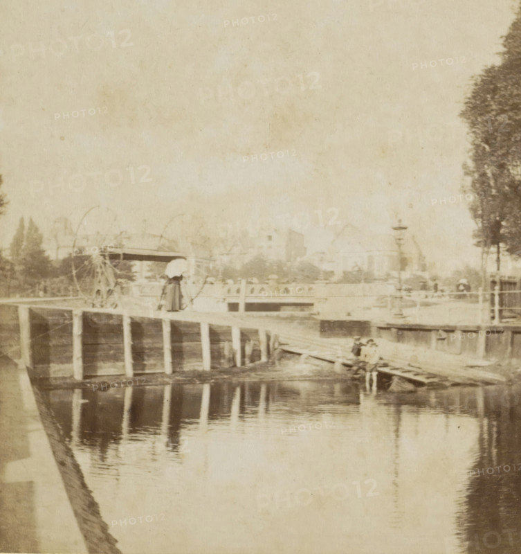View of the Ruysdaelkade in Amsterdam, Ruysdaëlkade, Amsterdam, stereograph, photographer: JG Beers, publisher: Nederlandsche Stereoscoop Maatschappij, 1873 - 1890, pasteboard, paper, albumen print, albumendruk, height 87 mm x width 179 mm, canals, waters (in city), pier, quay, wharf, Ruysdaelkade, A faded sepia scene showing a wooden pier structure extending over calm water with vertical supports reflected in the water, a figure standing on the pier wearing a long dress and a bonnet while holding an umbrella, another small group of figures sitting on a ramp near the water, distant blurred buildings and trees along the shoreline, a large tree silhouette on the right edge, and faint outlines of a ferris-wheel-like structure behind the pier.
