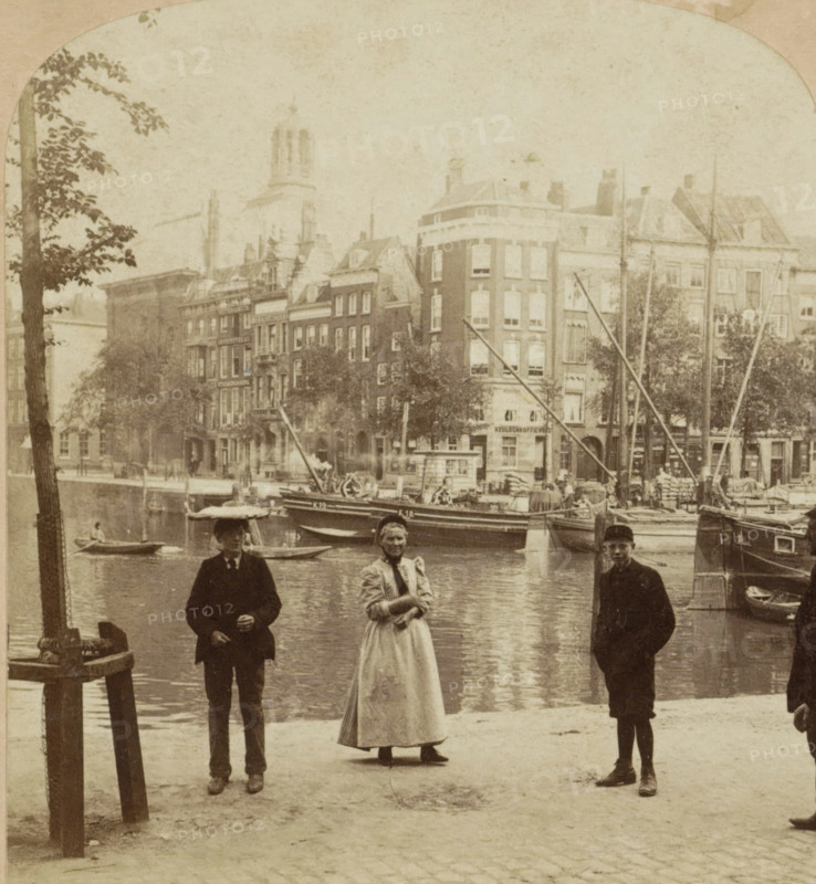 Holland. Rotterdam. Across the Canal, View of the Blaak in Rotterdam with the Lutheran church in the background, seen from the Schiedamsedijk, stereograph, number: '14691', retailer's mark: 'Sold only by Griffith & Griffith, PHILADELPHIA, CHICAGO, LONDON, HAMBURG, GER. MILAAN, ITALY.', Rotterdam, publisher: William Herman Rau, Philadelphia, 1896 - 1910, William Herman Rau, photographer, publisher, North America, pasteboard, paper, albumen print, height 89 mm x width 177 mm, urban housing, city-view in general, 'veduta', canals, waters (in city), Blaak, Schiedamsedijk, A waterfront scene with a row of tall buildings along the far bank, several moored boats with masts and rigging, and trees partially obscuring the facades; in the foreground three people stand on the quay: a woman wearing a long light-colored dress and apron with her hands clasped at her waist, flanked by two young men in dark clothing and caps standing with hands near their pockets; reflections of the boats and buildings...