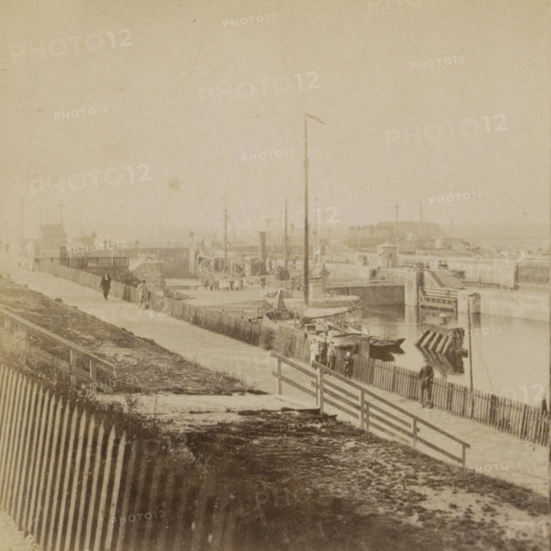 View of the harbor and locks of IJmuiden, IJmuiden, Collection A, Geographical sculpture gallery, collected by MJ Broekhuijsen, headmaster of a school in Amsterdam, stereograph, inscription: 'We are standing to the SE of the harbor. A boat is currently being locked through the lock.', number: 'A.5.', number: '5.', inscription: '58', publisher: Merkelbach & Co., Amsterdam, compiler (person in literature): Martien Johan Broekhuijsen, 1900 - 1902, photographer, compiler (person in literature), pasteboard, paper, albumen print, height 88 mm x width 177 mm, harbor, lift-lock, canal lock, A faded sepia scene showing a waterfront area with a boardwalk and piers, wooden fences running along a sloped embankment, small boats moored near the berths, tall masts and flagpoles rising from the docks, indistinct figures walking along the pathway and standing near the railings, a large pier structure extending into the water in the distance, and weathered ground and patterned shadows in the foreground.