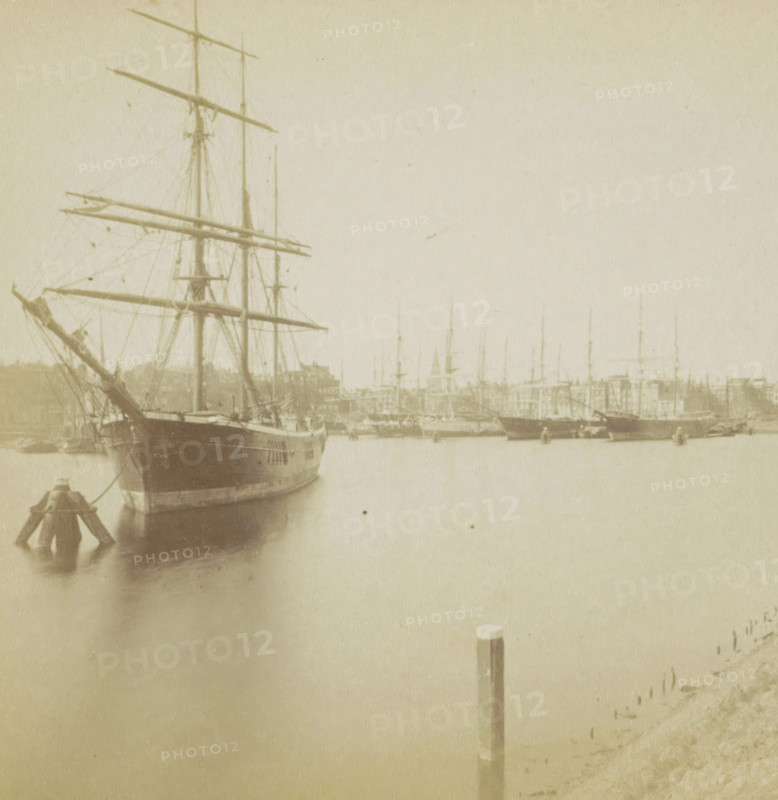 View of a city and a harbor with a ship in the foreground, Hollande, stereograph, Netherlands, 1873 - 1890, photographer, pasteboard, paper, albumen print, height 87 mm x width 177 mm, harbor, prospect of city, town panorama, silhouette of city, ships (in general), A faded sepia photograph showing a harbor scene with a large wooden sailing ship in the foreground at left, its tall masts, yards, and rigging visible, and the ship's bow facing slightly toward the viewer; a series of smaller masts and hulls of other vessels extend across the midground and recede into the distance, and a waterfront with low buildings is seen behind them; calm water fills the lower half of the image, reflecting the vessels, and a single wooden post rises from the water near the center-right; the overall image has a soft, aged texture and muted tones.