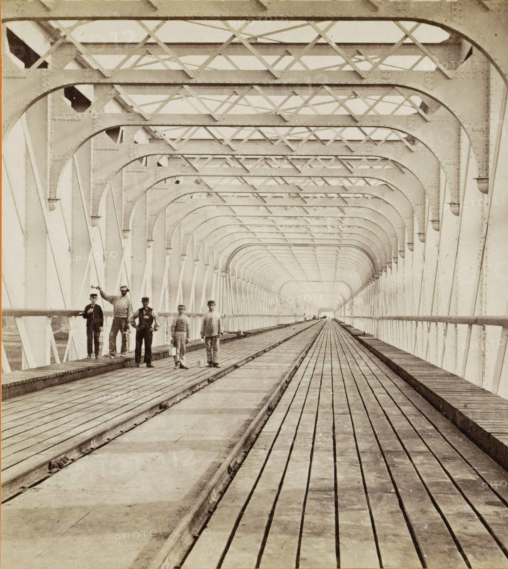 Kuilenburg railway bridge over the Lek, stereograph, photographer: Pieter Oosterhuis, Culemborg, before 1868-09-06, pasteboard, paper, albumen print, albumendruk, height 86 mm x width 176 mm, bridge in village across river, canal, etc. - II - ideal landscapes, Kuilenburg railway bridge, A long covered bridge interior with repeating metal arches and cross braces forming a tunnel-like perspective, wooden planks running lengthwise along the floor with a central track, railings along the sides, and a small group of men standing on the left side near the foreground wearing period clothing and hats.