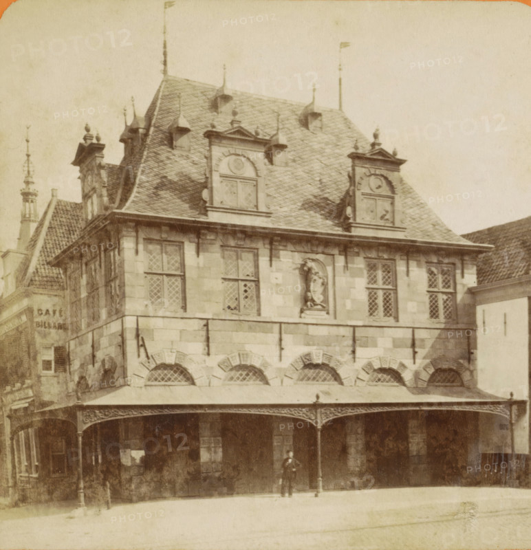 Horn. Roodenstein et le Waag, View of the Waag in Hoorn, Vues de Hollande, stereograph, inscription: '29', number: '1', Hoorn (North Holland), publisher: Léon & Lévy, Paris, publisher: J. Lévy & Cie, 1873 - 1890, photographer, pasteboard, paper, albumen print, height 87 mm x width 178 mm, weigh-house, Waag (Hoorn), A multi-story stone building with a steep tiled roof and decorative dormer windows occupies the image, featuring ornate gables, small finials along the roofline, and a statue set into a recessed niche on the upper facade; diamond-patterned window panes appear in rectangular windows, and a covered arcade with arched openings and ornate iron supports runs along the ground level, where a solitary figure stands beneath the canopy.