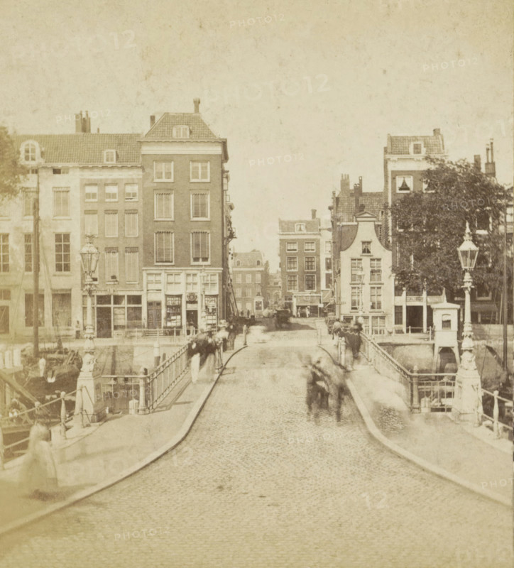 View of a bridge in Rotterdam, Leeuwen Bridge, Rotterdam, stereograph, inscription: '1200', 1861 - 1870, photographer, pasteboard, paper, albumen print, height 86 mm x width 175 mm, urban housing, bridge in city across river, canal, A cobblestone street runs from the foreground toward a bridge and a row of multi-story brick and plaster buildings, with tall narrow windows and sloped tiled roofs; ornate lamp posts and decorative railings line the bridge edges; indistinct blurred figures and horse-drawn carriages appear on the bridge and near the buildings; a large tree with dense foliage stands near one building on the right; canal water and moored boats are visible along the sides of the bridge; the overall image has a sepia tone and a slightly faded, aged appearance.