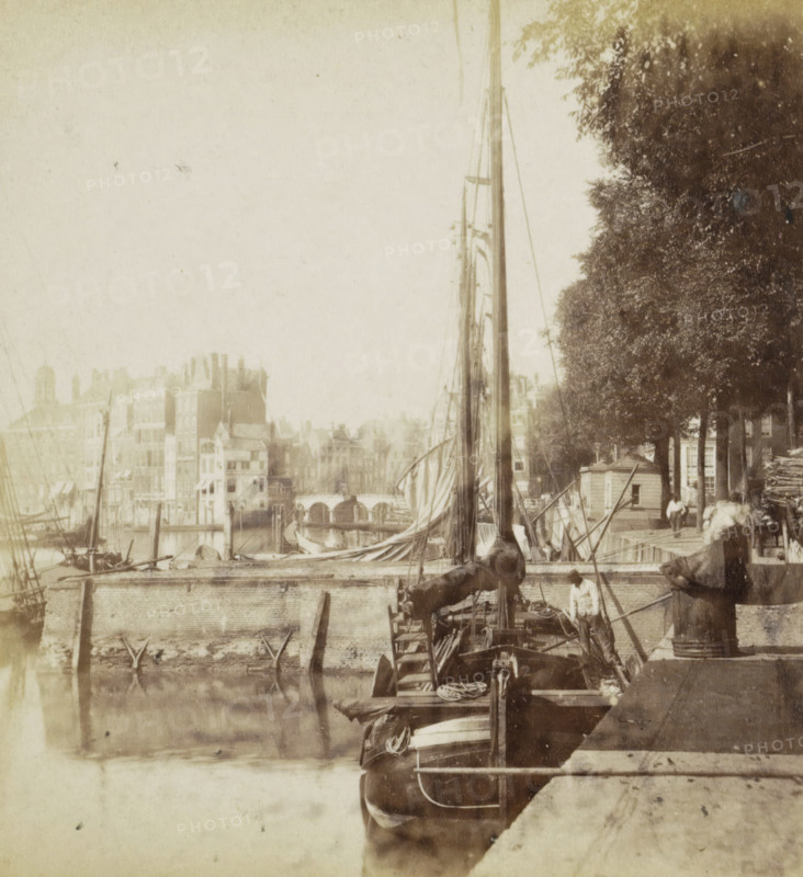 Cityscape with a harbor in Rotterdam, stereograph, Rotterdam, 1861 - 1870, photographer, pasteboard, paper, albumen print, height 75 mm x width 150 mm, harbor, city-view in general, 'veduta', ships (in general), A riverside scene with boats moored along a stone quay, tall masts and rigging rising above a vessel with rolled sails, a figure standing on one of the boats near the stern, another figure seated on the quay edge, clustered buildings with varied rooflines visible across the water, an arched bridge in the distance, and a row of leafy trees lining the quay casting dappled shade.