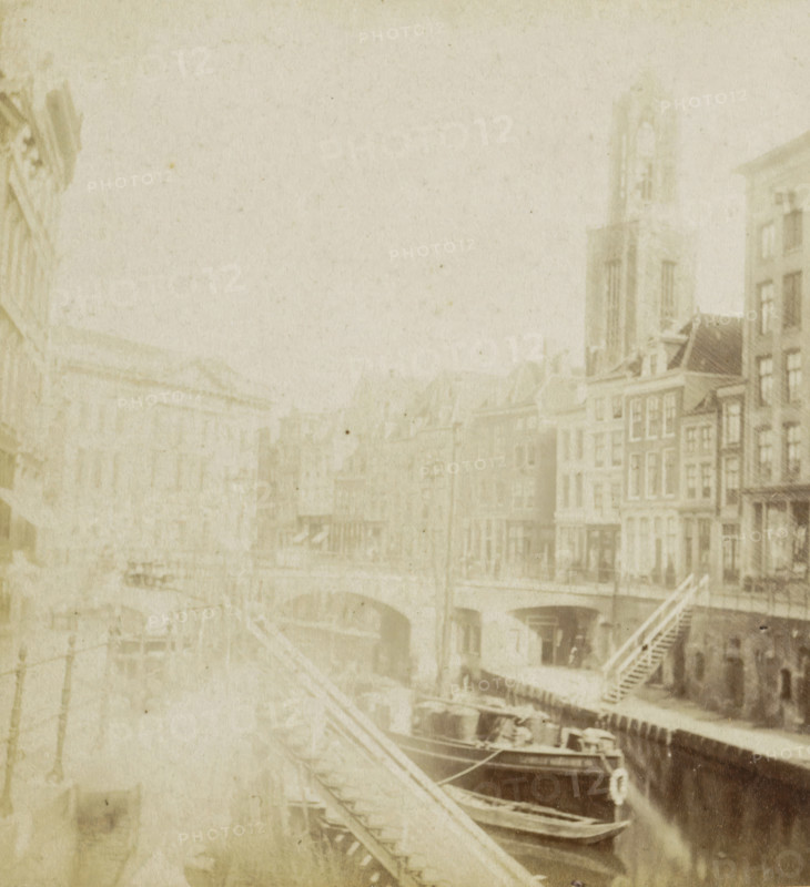 The Old Canal., Canal dit Oude Gracht., Der alte Kanal., The old Canal, View of the Oudegracht in Utrecht, Utrecht, stereograph, Rotterdam, 1873 - 1890, photographer, pasteboard, paper, albumen print, albumendruk, height 88 mm x width 179 mm, city-view in general, 'veduta', canals, waters (in city), parts of church exterior and annexes: tower, Oude Gracht (Utrecht), A faded sepia-toned scene shows a canal edged by multi-story buildings with narrow façades and peaked roofs, a tall tower rising among them, arched bridges crossing the water, wooden staircases leading down to the canal, several moored boats with visible hulls and rigging, and faint details of windows and doors along the waterfront, all appearing softened and washed by age.