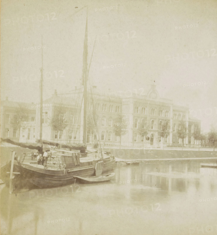 View of the Academic Hospital on the Catharijnesingel in Utrecht, The City Hospital., L'hopital., Das Krankenhaus., The Hospital, Utrecht, stereograph, inscription, 1871 - 1890, photographer, pasteboard, paper, albumen print, height 88 mm x width 178 mm, canals, waters (in city), hospital, ships (in general), Catharijnesingel, A pale, faded photograph showing a waterfront scene with several moored boats in the foreground, masts rising vertically and rigging visible, a small dinghy tied alongside, and the water reflecting the vessels; behind the boats a multi-story stone building with arched windows, decorative cornices, and a central pediment is visible across a paved area lined with small trees.