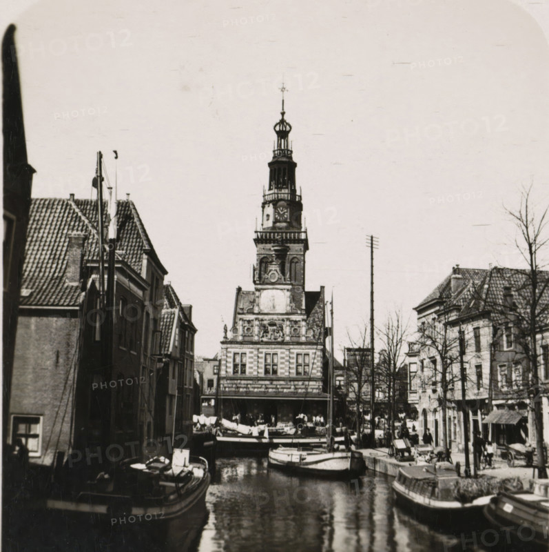 View of the Waag in Alkmaar, De Waag, Die Stadwage, The weight of the town Alkmaar, La balance de la ville Alkmaar, Alkmaar, stereograph, number: 'No. 2662.', number: 'R. 2662', 1900 - 1935, photographer, paper, gelatin silver print, developing gelatin silver print, height 89 mm x width 180 mm, city-view in general, 'veduta', weigh-house, Waag (Alkmaar), A narrow canal runs toward a multi-story building with an ornate clock tower rising above the rooftops, the tower topped by an openwork lantern and spire; moored boats fill the canal in the foreground with reflections on the water; rows of gabled buildings line both sides of the canal with tiled roofs and narrow facades; people are visible along the quays and near market stalls, and leafless trees stand between the buildings on the right side of the scene.