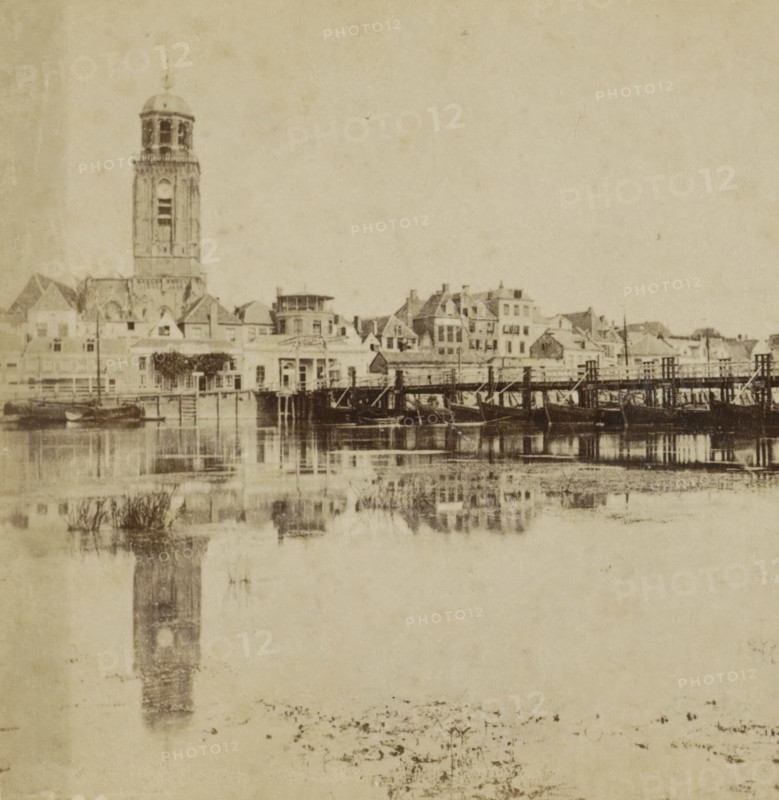 Deventer, View of the IJssel, the pontoon bridge and the Grote or Lebuïnus Church in Deventer, Collection A, Geographical sculpture gallery, collected by MJ Broekhuijsen, headmaster of a school in Amsterdam, stereograph, inscription: 'seen from the opposite side of the IJssel. Ship bridge, with a drawbridge on the city side.', number: '20.', publisher: Merkelbach & Co., Amsterdam, compiler (person in literature): Martien Johan Broekhuijsen, 1900 - 1902, photographer, compiler (person in literature), pasteboard, paper, albumen print, height 86 mm x width 176 mm, city-view in general, 'veduta', parts of church exterior and annexes: tower, pontoon bridge, Grote or Lebuinuskerk, A sepia-toned waterfront scene showing a row of buildings and houses along a shoreline with a tall tower rising among them, the tower featuring a domed top and an open belfry area, and smaller peaked and flat roofs surrounding it; a wooden pier or bridge extends across the water in front of the buildings with boats...