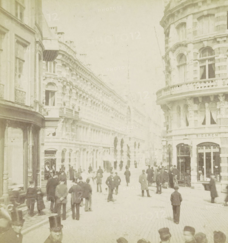 View of the Korte Poten in The Hague, The Hague, Korte Poten, stereograph, 1861 - 1870, photographer, pasteboard, paper, albumen print, albumendruk, height 88 mm x width 180 mm, urban housing, street, A faded street scene showing tall ornate buildings with arched windows and decorative facades lining both sides of the thoroughfare, a rounded corner building with a balustraded balcony visible on the right, shopfronts and window displays at street level, numerous people walking and standing along the street wearing long coats and hats of various styles, some clustered near shop entrances and others scattered down the roadway, a few figures nearer the foreground with top hats, a row of narrow dark trees or tall shrubs receding into the distance along the center of the street, and a pale sky above.