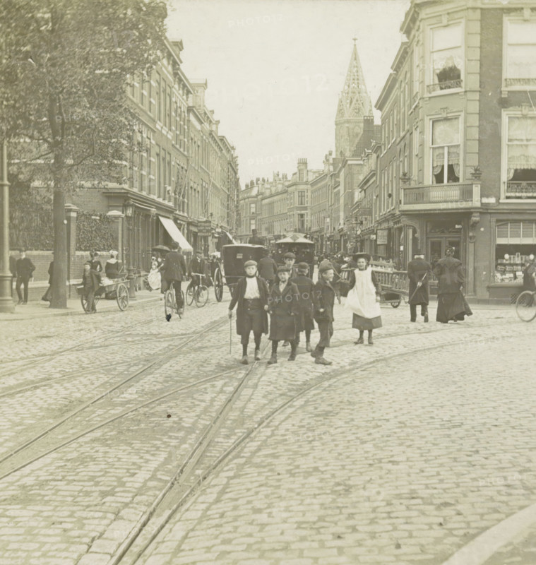s Gravenhage.- Prinsenstraat, View of the Prinsestraat in The Hague, stereograph, number: '72.', The Hague, 1882 - 1920, photographer, paper, pasteboard, gelatin silver print, developing gelatin silver print, height 87 mm x width 177 mm, urban housing, street, Prinsestraat, A cobblestone street with visible tram tracks runs toward a distant church spire flanked by rows of multi-story brick buildings; people in period clothing walk and stand in the street, including a group of boys in the foreground and a woman in an apron; horse-drawn carriages and a bicycle appear among the pedestrians; shopfronts and awnings line the buildings, some with window curtains and small balconies; a large tree stands on the left sidewalk and a handcart is visible near the curb.