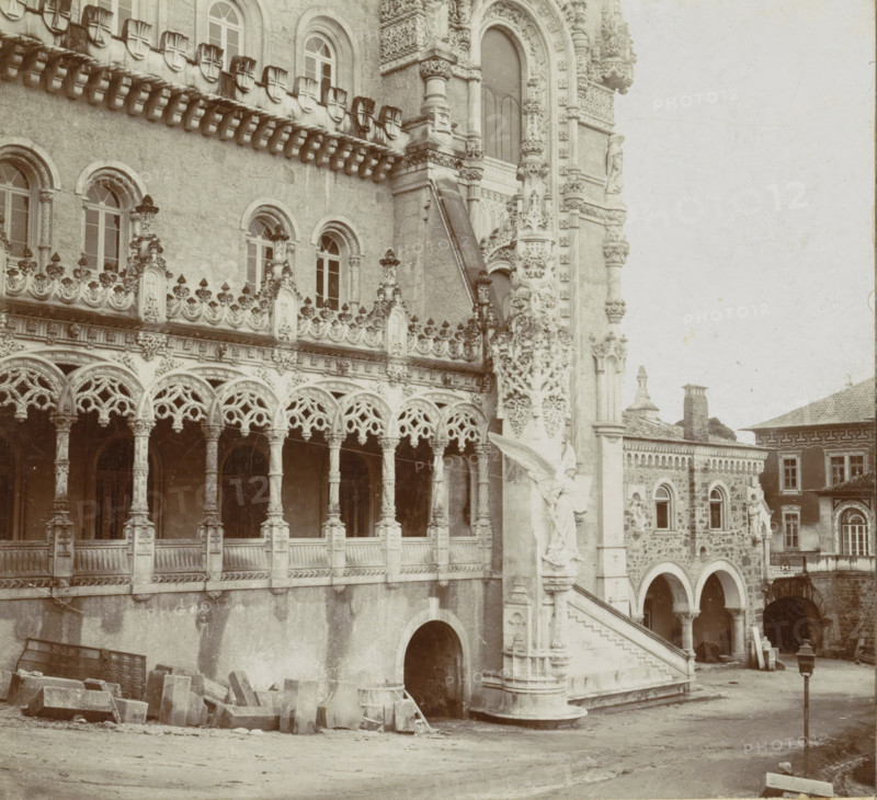 Domaine Royale du Bussaco. Façade principale de l'Hôtel, Facade of the Palácio Hotel do Buçaco, stereograph, inscription: 'Photographia Conimbricense', photographer: José Maria dos Santos, Palácio Hotel do Buçaco, 1869 - 1900, José Maria dos Santos, photographer, paper, pasteboard, baryta paper, gelatin silver print, developing gelatin silver print, height 88 mm x width 178 mm, palace, A detailed stone building facade with ornate carved arches and columns, a row of arched windows with decorative frames, and a balustraded gallery featuring intricate lace-like stonework; a sculpted stairway with carved figures and reliefs leads up beside a column richly decorated with floral and geometric motifs, while lower arches open into shadowed passageways and scattered stone blocks and masonry lie on the ground in front; adjoining structures with patterned stone walls, arched openings, and rectangular windows are visible at the side, and a lone lamp post stands in the courtyard area.