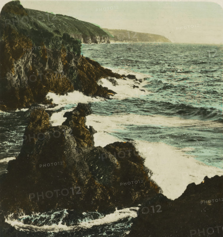Bornholm Löwenklikken, Kamelhovederne og Løvehovederne, rocks in the shape of camel and lion heads, Bornholm, stereograph, monogram: 'BW', number: 'S 24984', inscription: 'Dänemark. Bornholm. Löwenklippen., Denmark. Bornholm. Lion-cliffs., Denmark. Bornholm. Ecueils de lion., Dinamarca. Bornholm. Escollos de león.', photographer: BW, 1898 - 1935, paper, baryta paper, gelatin silver print, hand coloring, developing gelatin silver print, height 89 mm x width 168 mm, rock-formations, A rocky coastline with dark jagged cliffs extending into a choppy sea, waves breaking against the rocks and creating white foamy surf, the water showing variations of blue and green, a distant headland visible under a pale sky, and textured rock faces with patches of lighter and darker tones.