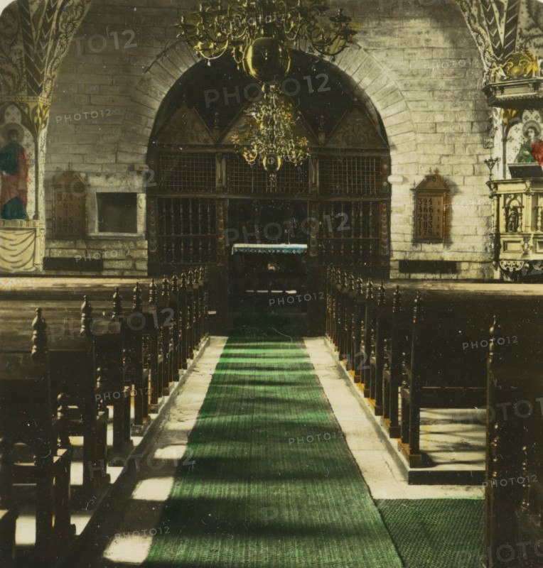 Interior of Husaby kyrka, the church of Husaby, Sweden. Kinekulle, 1000 jährige Kirche, stereograph, inscription: 'Schweden. Kinekulle. 1000jährige Kirche., Sweden. Kinekulle. Church 1000 years old., Suede. Kinekulle. Eglise de 1000 ans., Suecia. Kinekulle. Iglesia de 1000 años.', monogram: 'BW', number: '4', photographer: BW, Husaby, 1898 - 1935, paper, baryta paper, gelatin silver print, hand coloring, developing gelatin silver print, height 90 mm x width 170 mm, interior of church, A long central aisle with a green runner leads toward an arched stone entry and a dark wooden screen, flanked by rows of wooden pews with turned finials; above the screen hangs an ornate chandelier with a large central globe and decorative arms, and the surrounding stone walls show painted decorative panels and framed plaques; carved wooden and painted architectural details appear on the side altars, and the floor alongside the runner alternates between stone and shadowed areas.