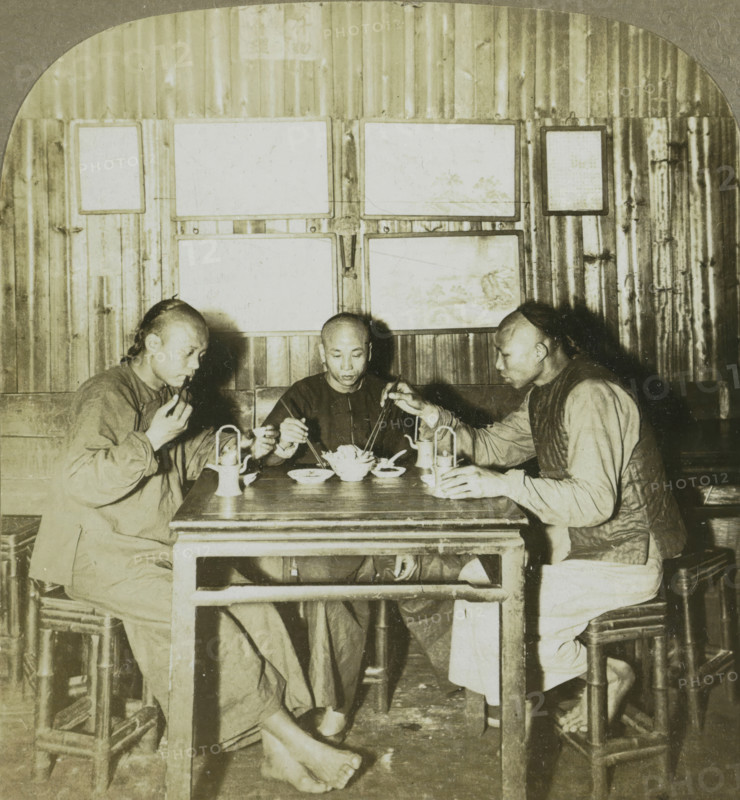Chinamen eating chow in a restaurant. Canton, China, Three Chinese men at a meal, stereograph, number: '168', copyright notice: 'Copyright 1902 by CH Graves', number: '2', photographer: Carleton Harlow Graves, Guangzhou, publisher: Universal Photo Art Company, Philadelphia (possibly), 1902, North America, paper, pasteboard, baryta paper, gelatin silver print, developing gelatin silver print, height 89 mm x width 178 mm, eating, Three men sit around a wooden table in a dim interior, each holding chopsticks and reaching toward a central bowl of food; the man on the left is turned slightly toward the center, wearing a loose garment and holding food near his mouth, the man in the middle faces forward with a neutral expression and a simple dark shirt, and the man on the right leans in from the side wearing a vest over a long-sleeved shirt. The table holds bowls, small plates, a teapot, and two lanterns, and the men's feet are visible beneath the table, one pair bare and resting on the floor...