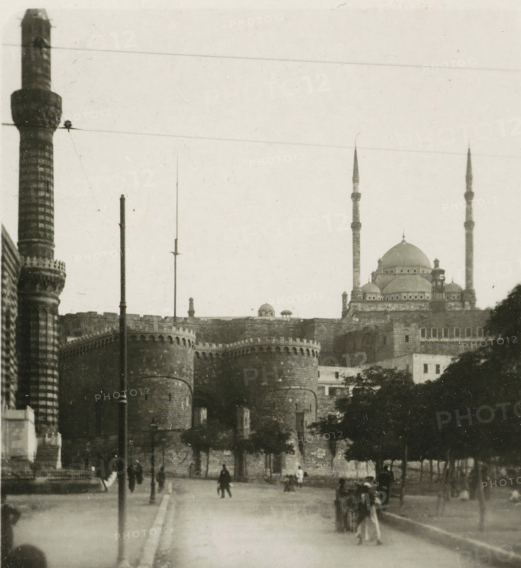 Port Said, View of a square in Port Said, stereograph, 1911, photographer, North Africa, paper, pasteboard, baryta paper, daylight gelatin silver print, height 90 mm x width 179 mm, square, circus, A monochrome scene showing a wide street leading toward a large stone fortress with rounded towers and crenellated walls, flanked on the left by a tall patterned column and on the right by a domed building with slender minarets rising above it; several vertical poles and wires cross the sky, trees line the right side of the street, and small groups of pedestrians and a few figures walking or standing are visible along the roadway.