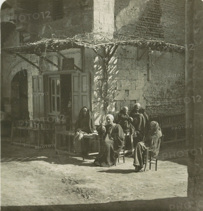 Luksor. Arabs Cafe, Guests in front of a café in Luxor, stereograph, number: '148', Luxor, publisher: Neue Photographische Gesellschaft, Steglitz, 1909 - 1935, photographer, North Africa, paper, baryta paper, gelatin silver print, developing gelatin silver print, height 88 mm x width 179 mm, pavement, terrace in front or inn or restaurant, An exterior scene shows a weathered stone building with a wooden door and shutters partly open beneath a simple pergola of branches and vines; in front of the building a group of people wearing dark, draped garments and head coverings sit and stand on benches and chairs, some leaning forward and some with arms crossed, their faces partially in shadow; the ground in front is bare earth with faint footprints and patches of darker material, and the sunlight casts irregular shadows from the pergola and nearby structures across the wall and ground.
