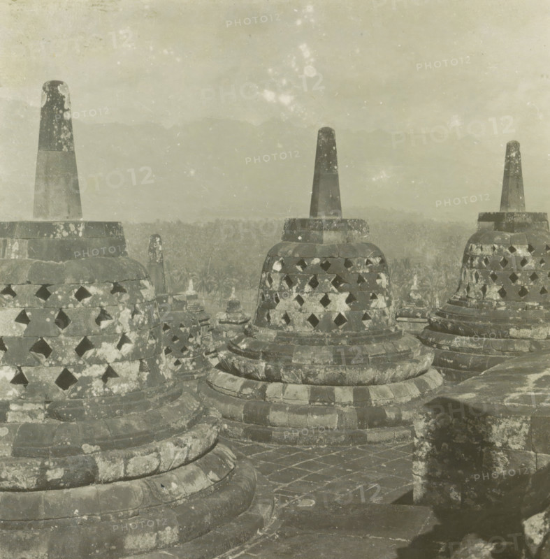 Stupas on Borobudur, Java, Boroboedoer, stereograph, Borobudur, 1900 - 1940, photographer, Central Java, paper, pasteboard, baryta paper, gelatin silver print, developing gelatin silver print, height 90 mm x width 185 mm, places and objects of worship, Hinduism, Buddhism, Jainism, Stone stupas with conical pinnacles and perforated bell-shaped bodies fill the foreground, their surfaces mottled with lichen-like patches and weathering; layered circular bases and stone walkways with visible joints and wear surround them, and a hazy distant landscape of treetops and faint mountain silhouettes recedes under a pale, lightly speckled sky.