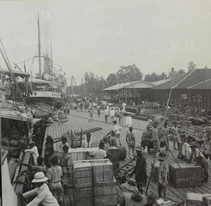 View of a harbor in Indonesia, stereograph, inscription: 'B' Massin', Indonesia, 1890 - 1930, photographer, paper, pasteboard, baryta paper, daylight gelatin silver print, height 87 mm x width 178 mm, harbour, A busy wooden pier filled with people carrying and moving large crates and bundles, with several men wearing hats and loose clothing and some women among them, a large multi-deck ship docked to the left showing lifeboats, rigging, and tall masts, rows of warehouses with sloped roofs along the right side, stacks of pots and coiled ropes near the buildings, palm trees and other foliage behind the warehouses, wooden planks forming the pier surface, various handcarts and timber scattered across the scene, and groups of workers clustered and walking along the pier.