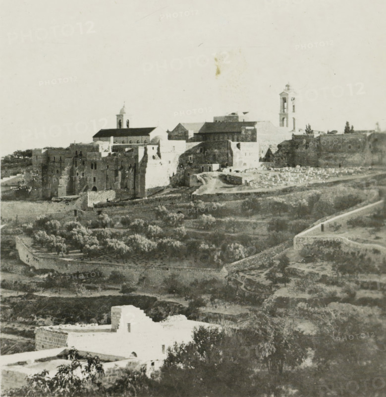 Bethlehem, View of Bethlehem, stereograph, address: 'Parklaan 49, HAARLEM, (Holland)', inscription: 'These stereo-photos are being offered for sale and sold wholesale and retail by the ........., ', number: '166.', monogram, inscription: 'Déposée.', photographer: Richard Sochor, publisher: De Wereld-Tourist, Haarlem, 1903, Palestine, paper, baryta paper, gelatin silver print, gelatin silver developing print, height 88 mm x width 179 mm, city-view in general, 'veduta', A wide panoramic view shows an old stone cityscape with a cluster of large masonry buildings and fortified walls on a hill, two prominent bell towers rising above the roofline, pitched roofs and flat terraces visible among the structures, and a road or path winding along the base of the walls. In the foreground there are terraced fields and groves with rows of trees separated by low stone walls, scattered smaller buildings with whitewashed facades, and irregular patches of bare earth and vegetation. The overall image is m...
