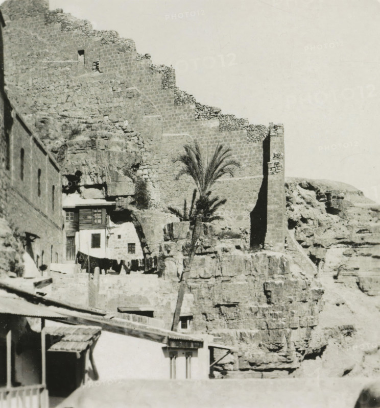 Vallée du Cédron. Le couvent Mar Saba, Mar Saba monastery in the Kidron Valley, Israel, stereograph, inscription: 'Déposée.', address: 'Parklaan 49, HAARLEM, (Holland)', number: '189.', inscription: 'These stereo photographs are marketed and sold wholesale and retail by the ........., ', monogram, photographer: Richard Sochor, Mar Saba, publisher: De Wereld-Tourist, Haarlem, 1903, Jerusalem, Palestine, Kidron-dal, paper, baryta paper, gelatin silver print, developing gelatin silver print, height 90 mm x width 178 mm, abbey, monastery, convent, Roman Catholic church, A stone fortress wall with stepped crenellations rises against a pale sky above a rocky cliff face, with rough stone blocks and rubble visible along the wall and cliff; near the base of the wall there is a tall narrow vertical shadowed opening and a slender palm tree with feathery fronds stands in front of the rock; adjacent to the cliff are small buildings with stone and plaster facades, wooden shutters, a balcony, and a l...