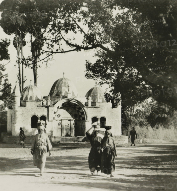 Jaffa. Fontaine Abou Nadoud, Sabil Abu Nabbut fountain in Jaffa, Israel, stereograph, address: 'Parklaan 49, HAARLEM, (Holland)', inscription: 'Déposée.', monogram, number: '91.', inscription: 'These stereo photographs are marketed and sold wholesale and retail by the ........., ', photographer: Richard Sochor, Sabil Abu Nabbut, publisher: De Wereld-Tourist, Haarlem, 1903, Ottoman Empire, Jaffa, paper, baryta paper, gelatin silver print, developing gelatin silver print, height 89 mm x width 180 mm, utilitarian, public pump or water fountain, A black and white photograph showing a stone building with domed roofs and an arched entrance partially framed by overhanging tree branches and foliage; several figures walk in the foreground, some carrying bowls or containers on their heads, and more people are visible near the building and in the shaded areas under the trees; the scene includes sunlit ground with shadows from the trees and architectural details like arches and small columns on th...
