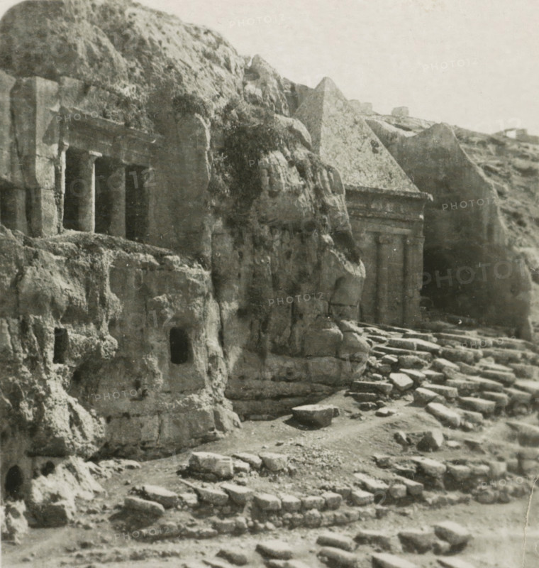Jerusalem Sépulcres St. Jacques et St. Zachari, Rock Tombs of Benei Hezir and St. Zacharia, Kidron Valley, stereograph, number: '159.', inscription: 'Déposée.', monogram, address: 'Parklaan 49, HAARLEM, (Holland)', inscription: 'These stereo photographs are marketed and sold wholesale and retail by the ........., ', photographer: Richard Sochor, Kidron-dal, publisher: De Wereld-Tourist, Haarlem, 1903, Jerusalem, paper, baryta paper, gelatin silver print, developing gelatin silver print, height 88 mm x width 179 mm, grave, tomb, Tomb of Benei Hezir, Tomb of Zechariah, A grayscale photograph of a rocky cliff face with several rectangular carved openings and recessed columns set into the rock, adjacent to a stepped stone pathway and scattered blocks; the rock surfaces show weathered textures and erosion, with a triangular rock formation above one carved recess and uneven rocky ledges and small cavities visible across the scene.