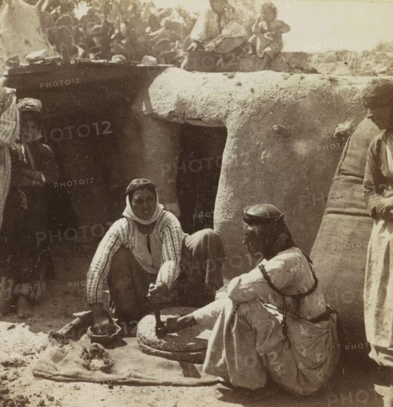 Frauen an der Mühle. (Nazareth), Women grinding grain in Nazareth, Collection Hentschel, stereograph, number: '813.', inscription: 'Déposé.', inscription: 'Registered.', Nazareth, publisher: Bruno Hentschel, Leipzig, 1905, photographer, Saxony, pasteboard, paper, albumen print, albumendruk, height 89 mm x width 175 mm, handicrafts, other plant material, A sepia-toned photograph showing a group of people seated and crouched outdoors beside a rough stone structure with an opening and a rounded overhang; two women in patterned garments and head coverings are prominent, one crouching and reaching toward a stone mortar with a pestle held by another seated woman in layered clothing, while a man with a headwrap sits behind them and another figure stands partially visible on the right wearing a striped textile draped over the shoulder; the ground is sandy with scattered textiles and a shallow bowl nearby, and the upper edge of the image shows additional figures and irregular stone or earth for...