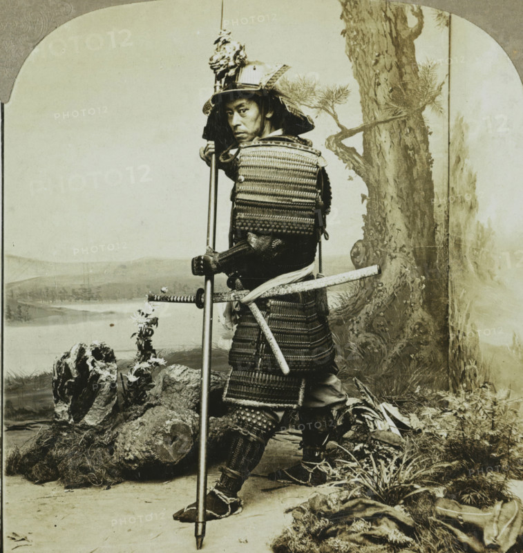 Ancient warrior costume of the Japanese. Japan, Japanese soldier in traditional dress, stereograph, copyright notice: 'Copyright 1902 by CH Graves.', number: '30', number: '59', number: '190.', photographer: T. Enami (possibly), Japan, publisher: Universal Photo Art Company, Philadelphia, 1900, North America, paper, pasteboard, baryta paper, gelatin silver print, developing gelatin silver print, height 89 mm x width 178 mm, the soldier, the soldier's life, A sepia-toned portrait of a person wearing layered armor and a decorated helmet, standing with a long pole held vertically and a sheathed sword at their side; the armor shows woven and plated textures with straps and lacing visible, and the helmet has an ornate crest and flared neck guard. The person looks toward the camera with a neutral facial expression, one hand gripping the pole and the other resting near the sword hilt. The scene includes a painted backdrop with a tree trunk and distant landscape, and the foreground shows rocks...