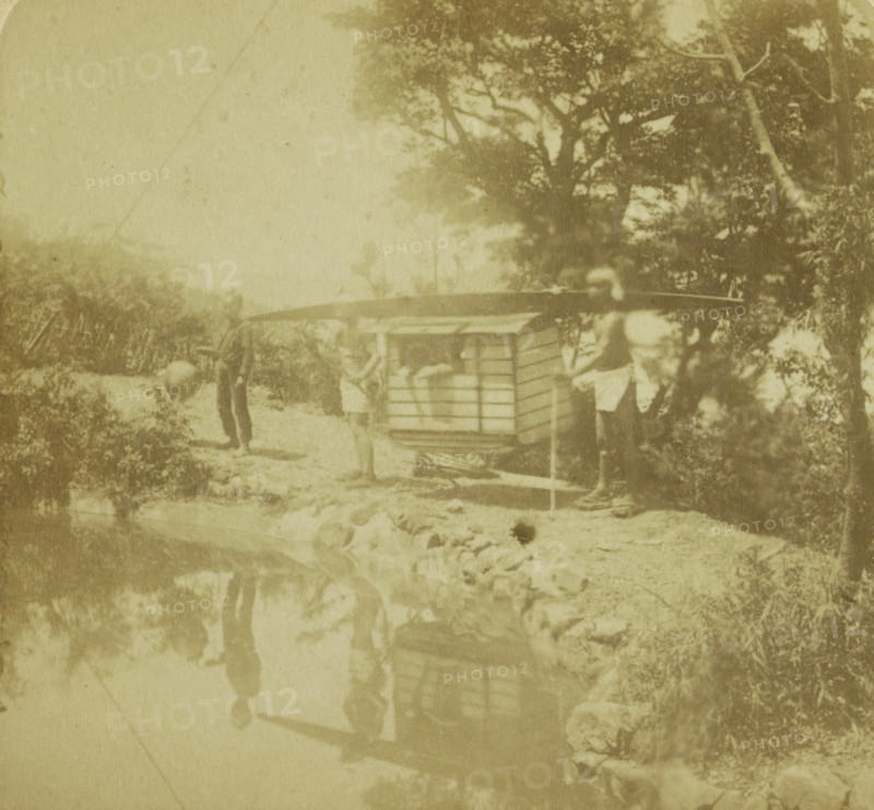 Travelers with servants in a tandem, Nagasaki, Japan, Traveling, Geographical gallery of images, collected by MJ Broekhuijsen, head of a school in Amsterdam, Collection H, stereograph, number: '23', inscription: 'from Nagasaki (see pl.7) into the interior. The front coolie carries, ', publisher: Merkelbach & Co., Amsterdam, compiler (person in literature): Martien Johan Broekhuijsen, 1900 - 1902, photographer, compiler (person in literature), Kyushu, pasteboard, paper, albumen print, height 89 mm x width 179 mm, passenger carriage, A faded sepia photograph showing a riverside scene with a wooden boxlike structure on a pole platform near the bank, a person standing beside it holding a long horizontal pole across their shoulders, another person nearby carrying a spherical object, vegetation and trees along the right side, and a reflective water surface in the foreground mirroring the figures and structures.